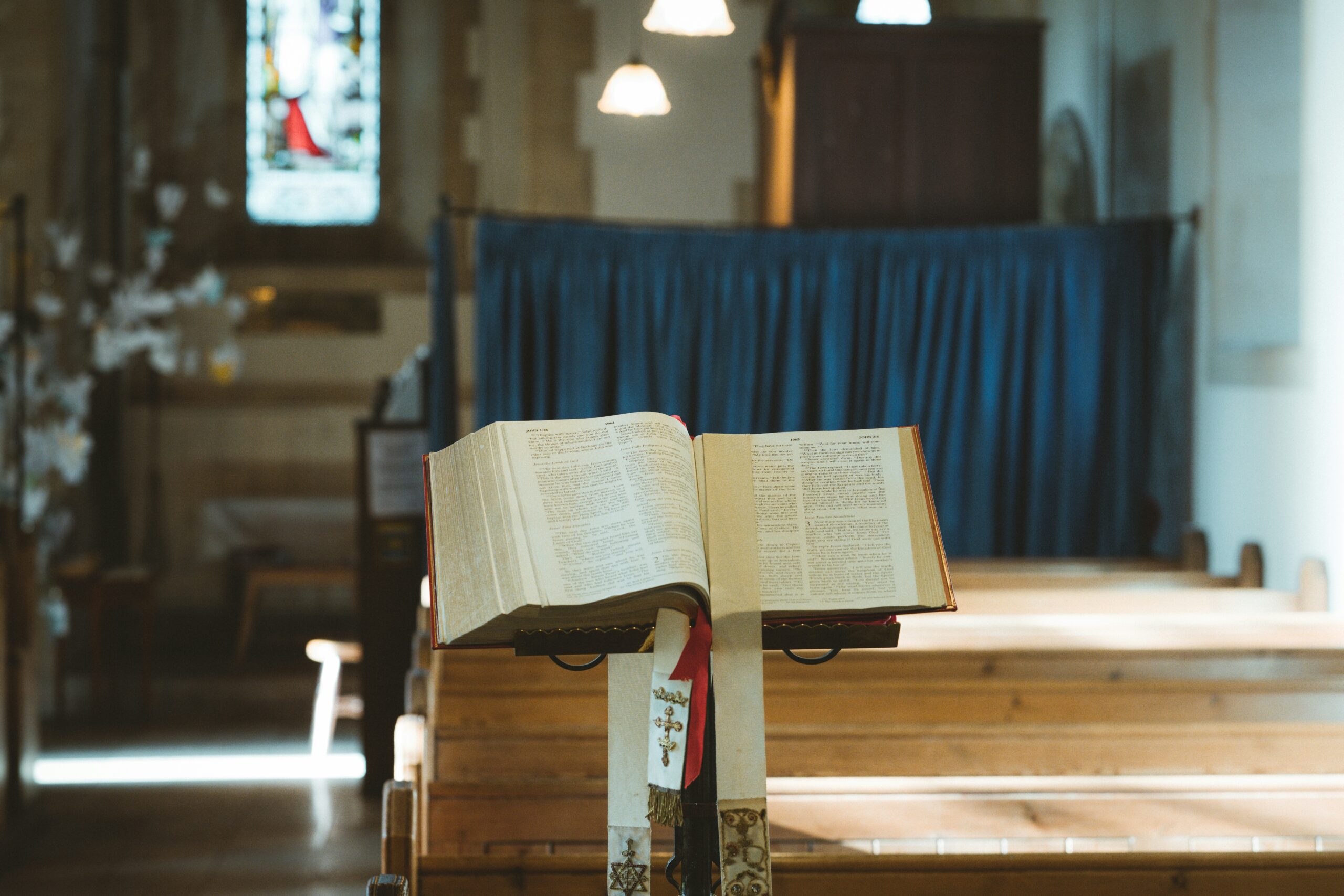 Close-up of an open Bible resting on a wooden lectern inside All Saints Church in Holbeach, with sunlight streaming through stained glass windows and illuminating the pages.